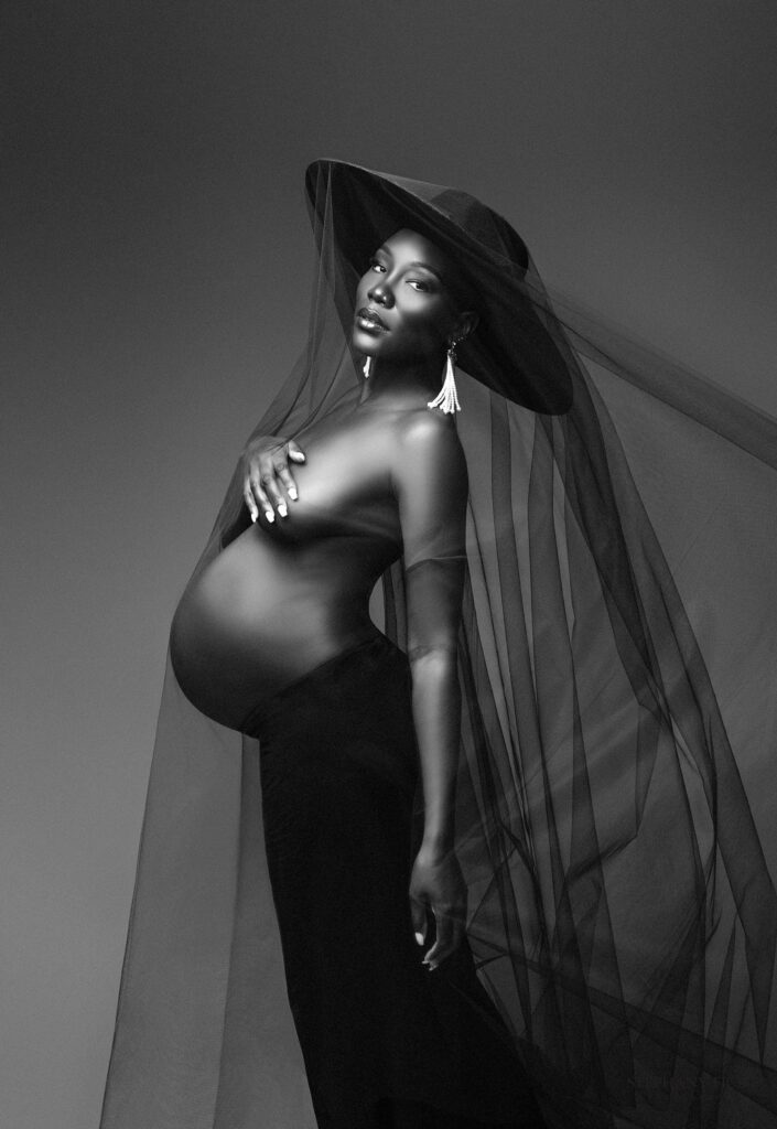Black and White Elegant maternity portrait in studio with mother in gown surrounded by soft props and warm lighting.