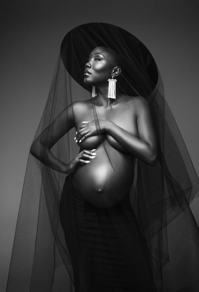 Black and White Elegant maternity portrait in studio with mother in gown surrounded by soft props and warm lighting.